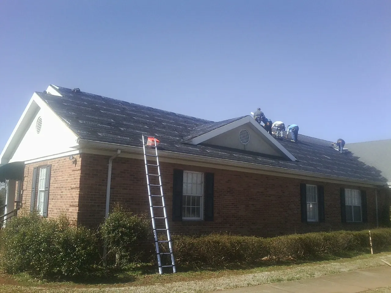 Crew tearing shingles off a brick ranch roof with a ladder set — Metal Roof Repair work by our crew in Malta