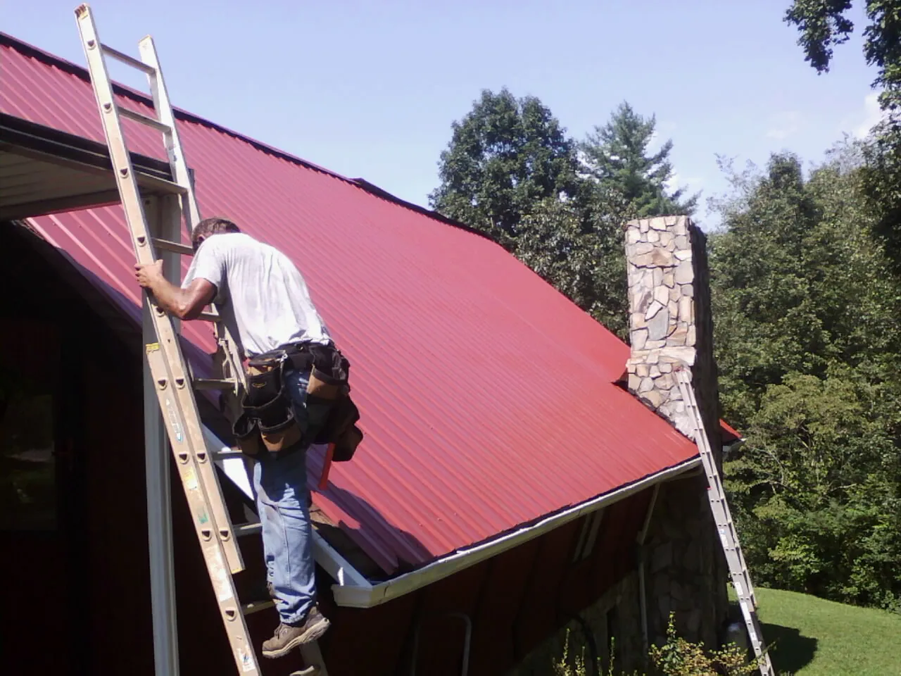 Roofer on a ladder beside a red metal roof and stone chimney — Soffit & Fascia Repair by Roofing Brothers in Malta